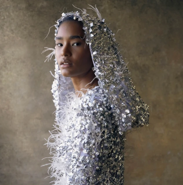 Striking portrait of a woman with fair skin in a silver beaded hood and feathered top, set against a neutral backdrop.