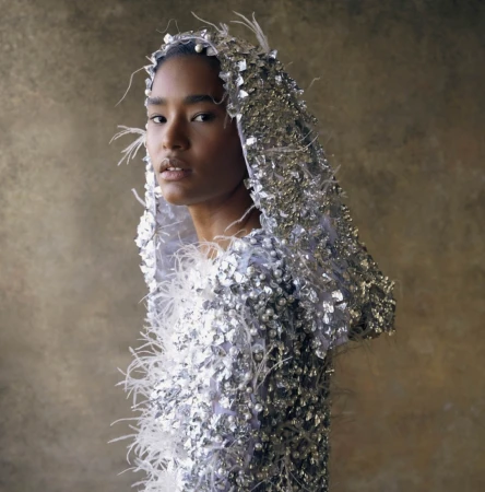 Striking portrait of a woman with fair skin in a silver beaded hood and feathered top, set against a neutral backdrop.