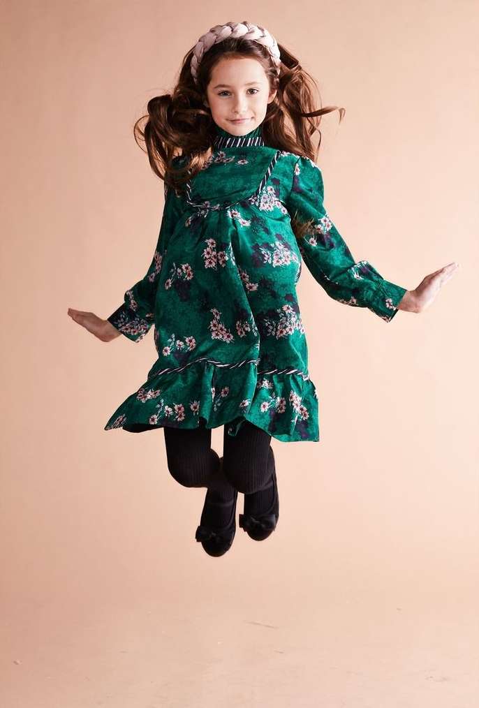 A young girl in a green floral dress and black tights jumps in front of a beige background, smiling with arms outstretched in the air.