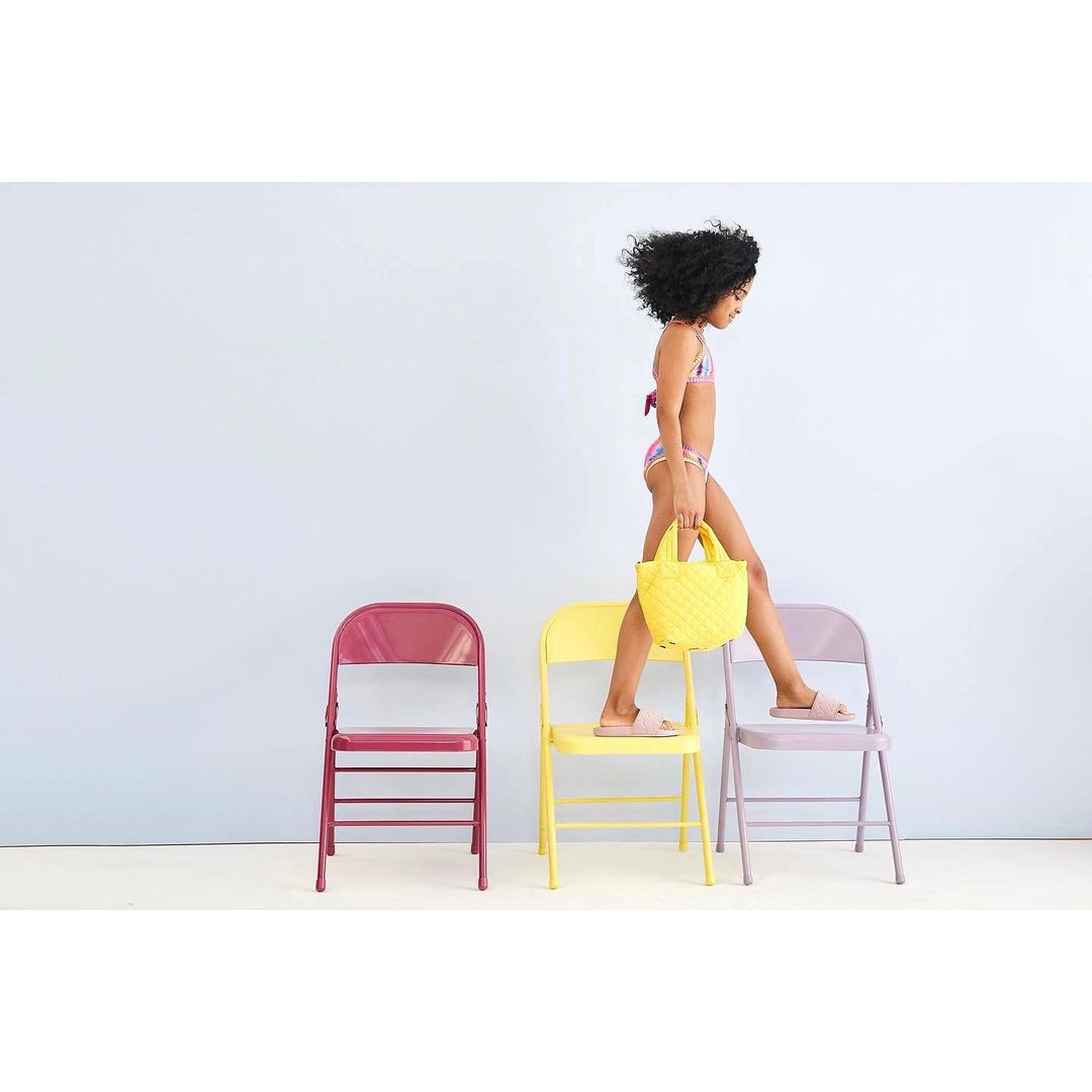 Young child in swimwear walks across three colorful folding chairs, carrying a yellow handbag against a pale blue background.
