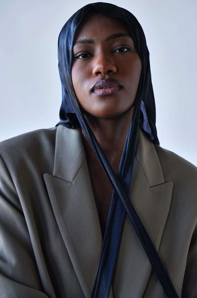 Portrait of a dark-skinned adult woman wearing a navy blue head covering and an oversized tan blazer against a neutral background.