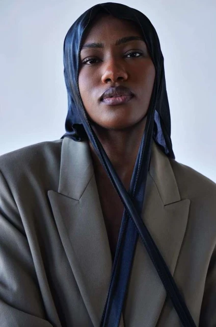 Portrait of a dark-skinned adult woman wearing a navy blue head covering and an oversized tan blazer against a neutral background.
