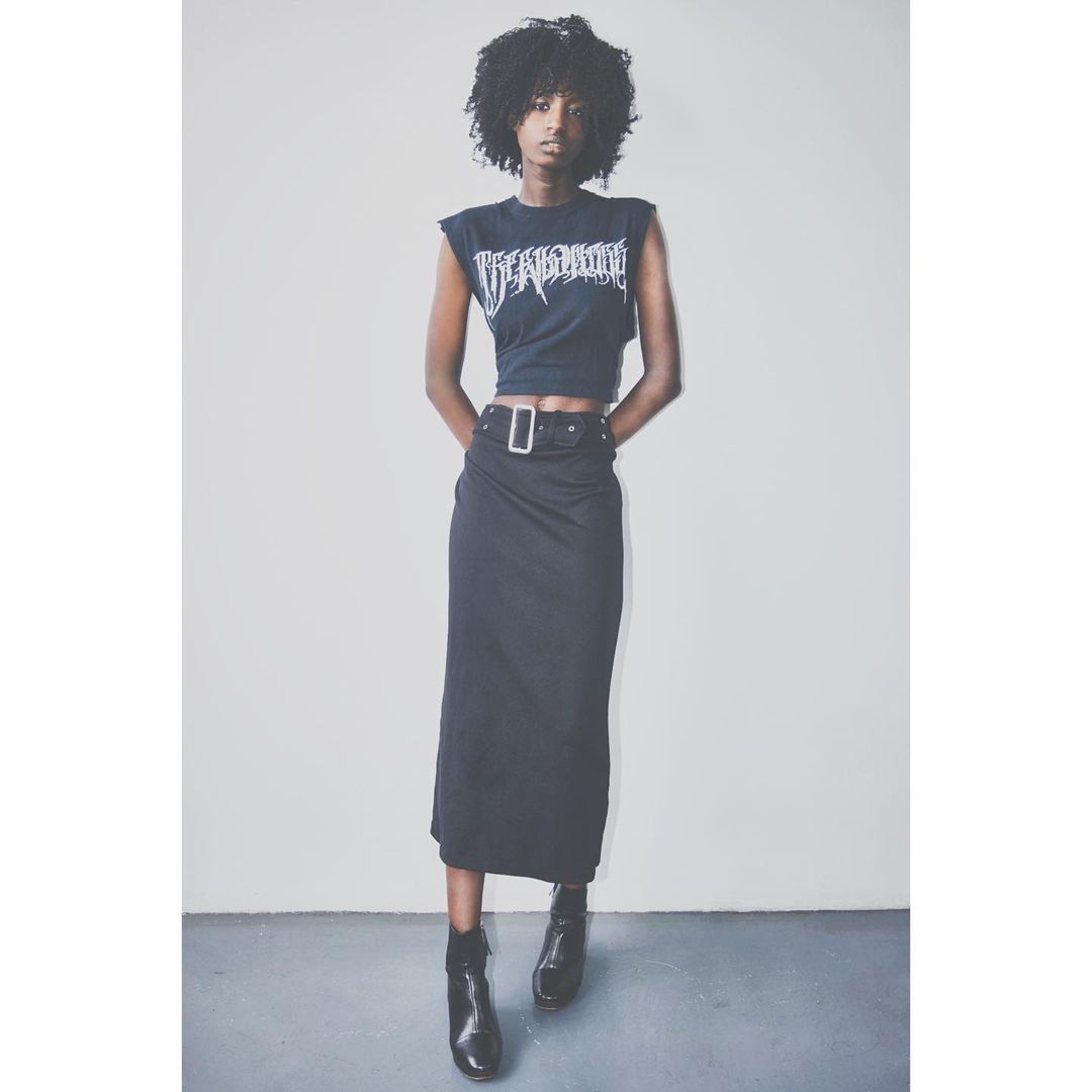 Woman in crop top and maxi skirt posing against grey studio background. She wears ankle boots, with a black belt and silver buckle.