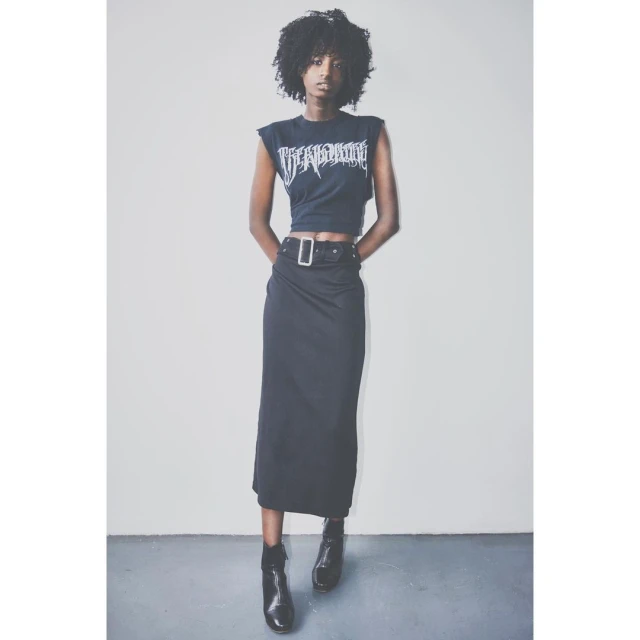 Woman in crop top and maxi skirt posing against grey studio background. She wears ankle boots, with a black belt and silver buckle.