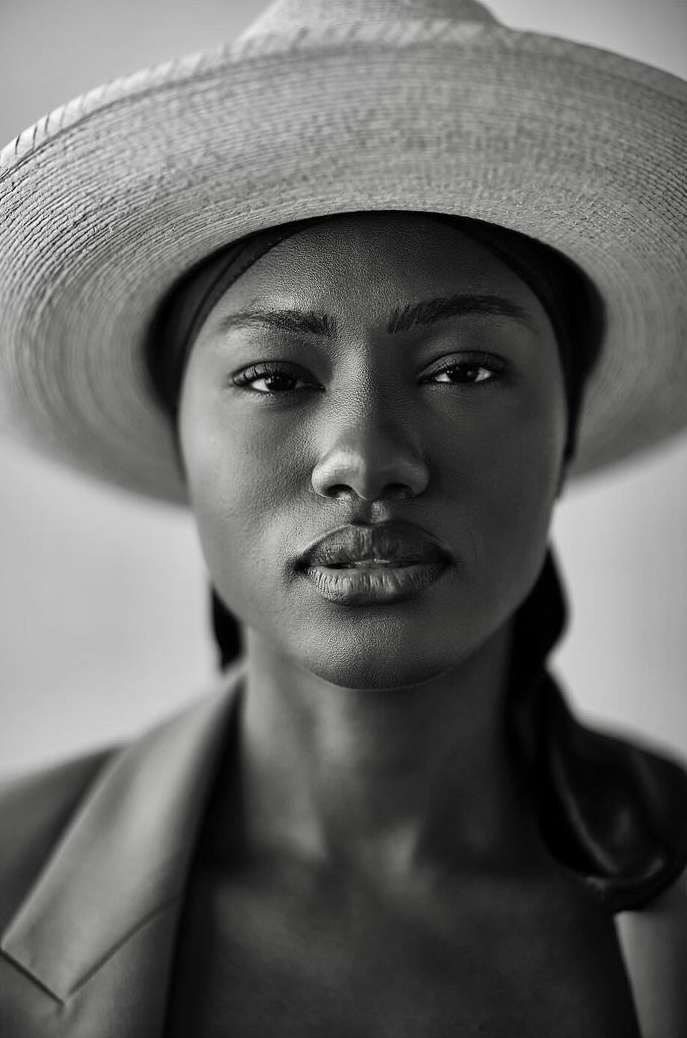 Striking black and white portrait of a woman in a blazer and straw hat, showcasing neutral facial expression and close-up framing.