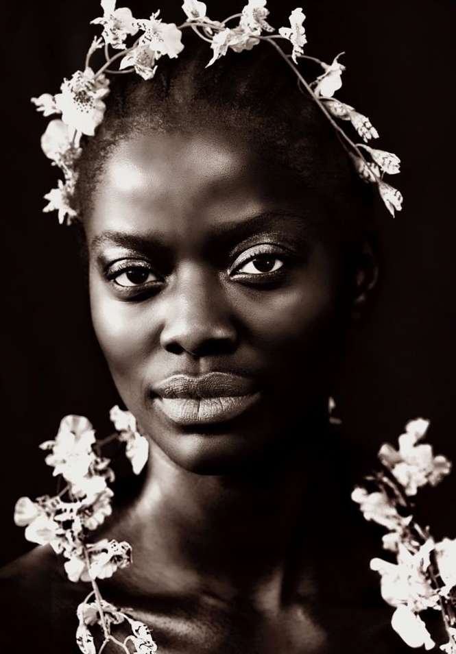 Portrait of a black woman wearing a floral headband and flower garlands against a dark background, photographed by Brian Sassmann.