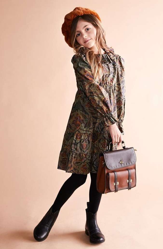 Young girl wearing a patterned dress, brown beret and carrying a leather satchel against a seamless beige backdrop.