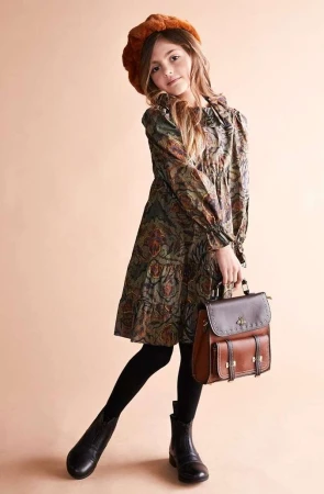 Young girl wearing a patterned dress, brown beret and carrying a leather satchel against a seamless beige backdrop.