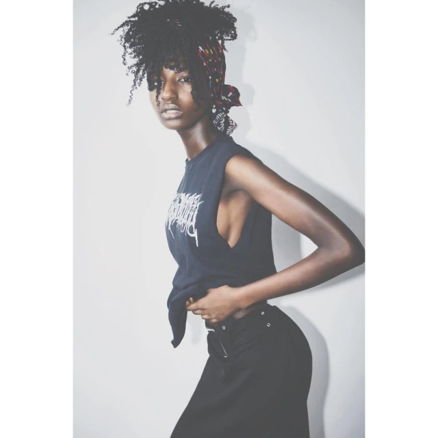 Young Black woman in a sleeveless graphic tee and headscarf poses against a white wall in a studio, looking confidently at the camera.