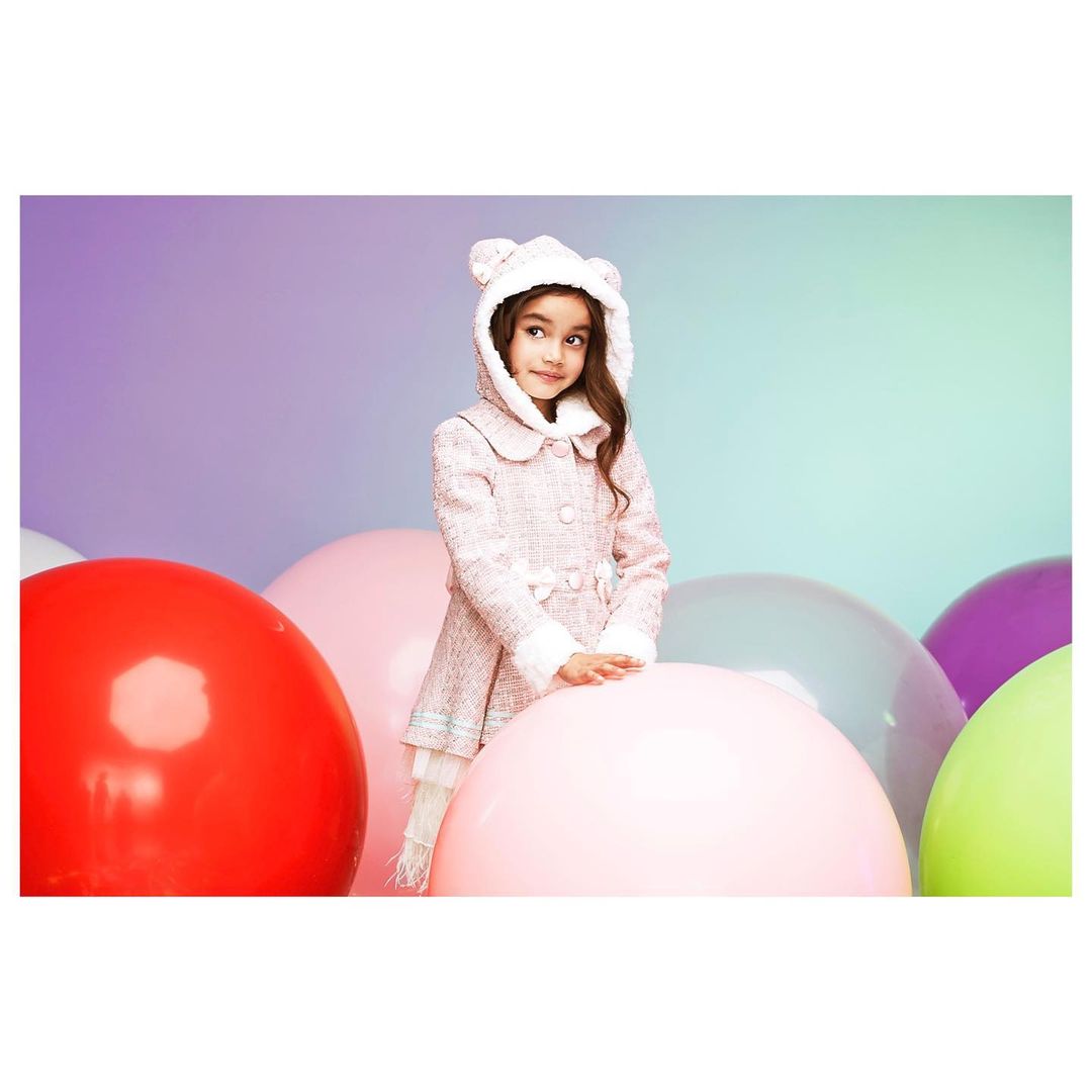 Young girl in pink hooded coat surrounded by large colorful balloons poses in a studio with a light pastel background.