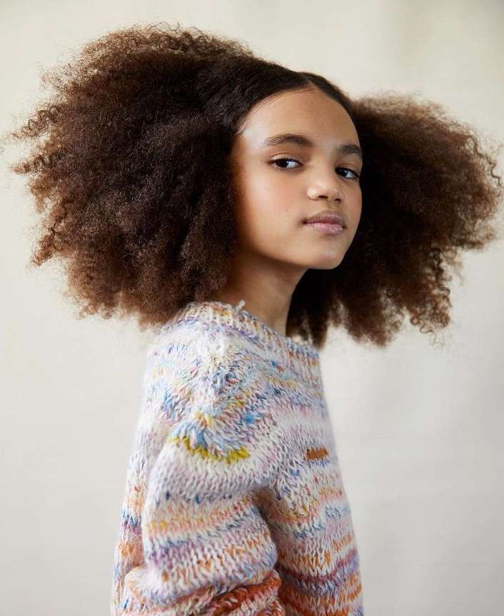 Portrait of a young African American girl with natural afro hair wearing a multicolored knit sweater against a neutral background.