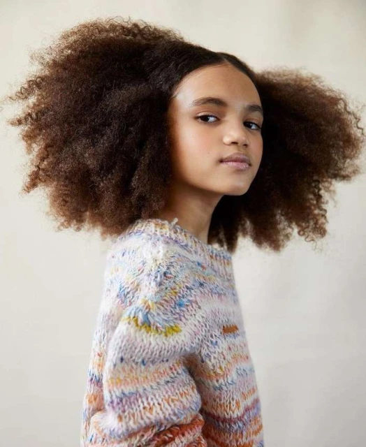 Portrait of a young African American girl with natural afro hair wearing a multicolored knit sweater against a neutral background.