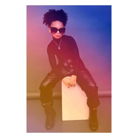 Young Black model wearing dark sunglasses, black turtleneck, and leather cargo pants posing on a light box.