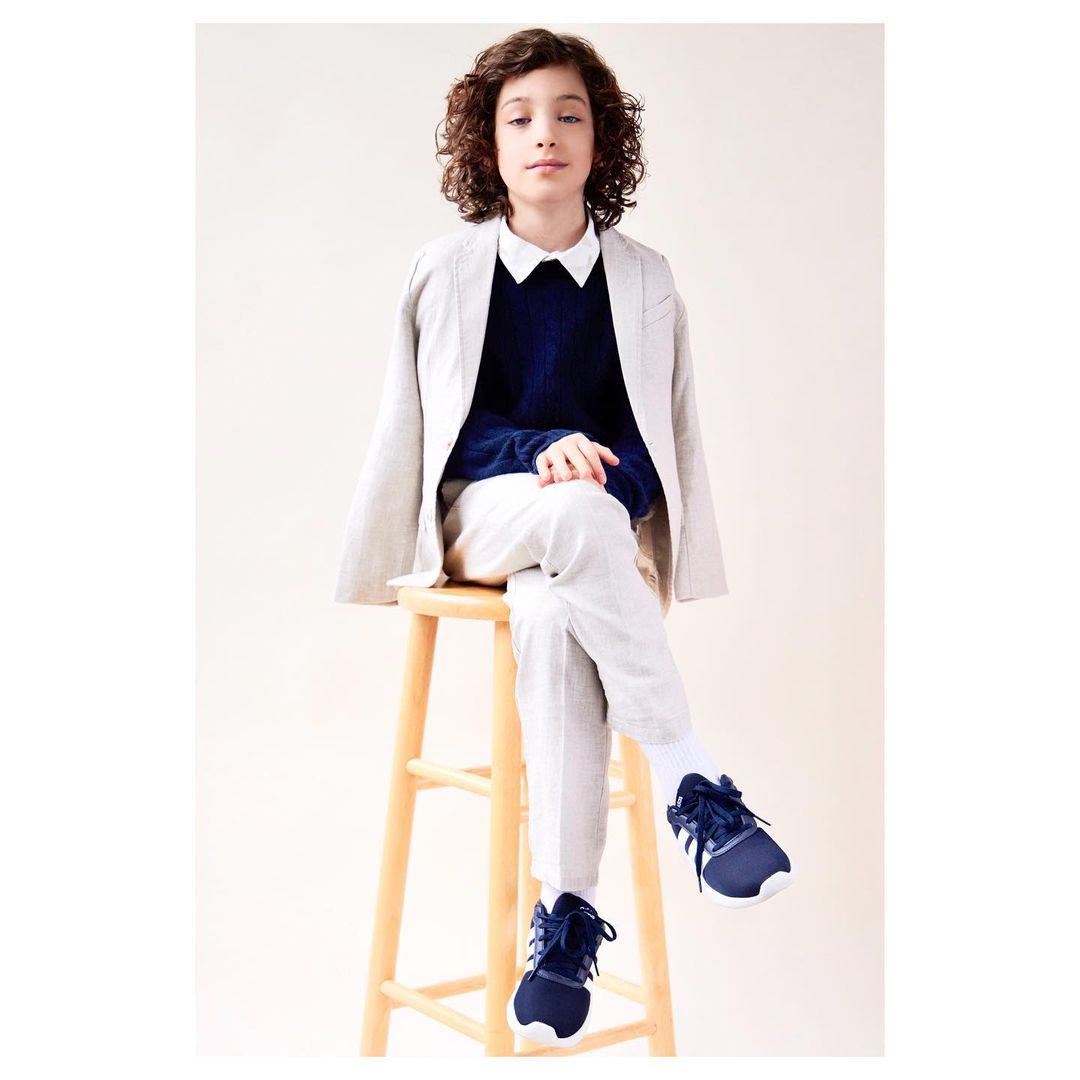 A child in a blazer and sweater sits on a stool in a studio, looking at the camera, with a neutral expression and light background.