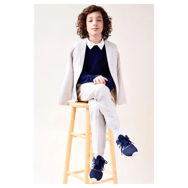 A child in a blazer and sweater sits on a stool in a studio, looking at the camera, with a neutral expression and light background.
