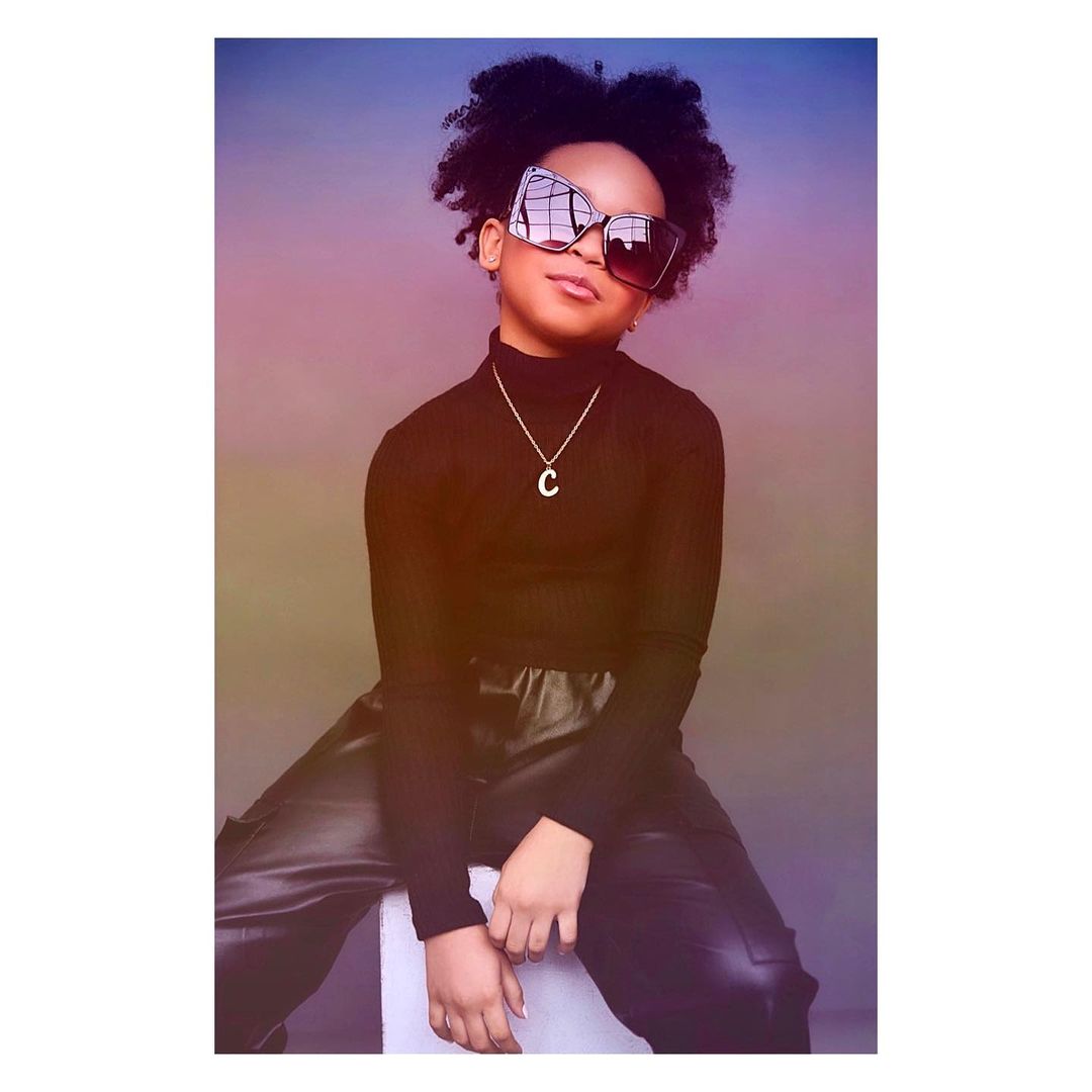 Stylish young girl with afro hair, wearing oversized sunglasses, a black turtleneck, a gold necklace, and leather pants, sitting indoors.
