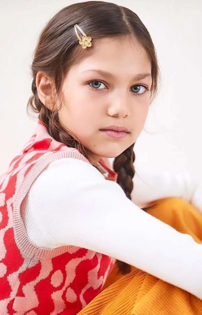 Portrait of a young girl with braided hair, wearing a patterned vest and a hair clip, sitting indoors with a neutral background.
