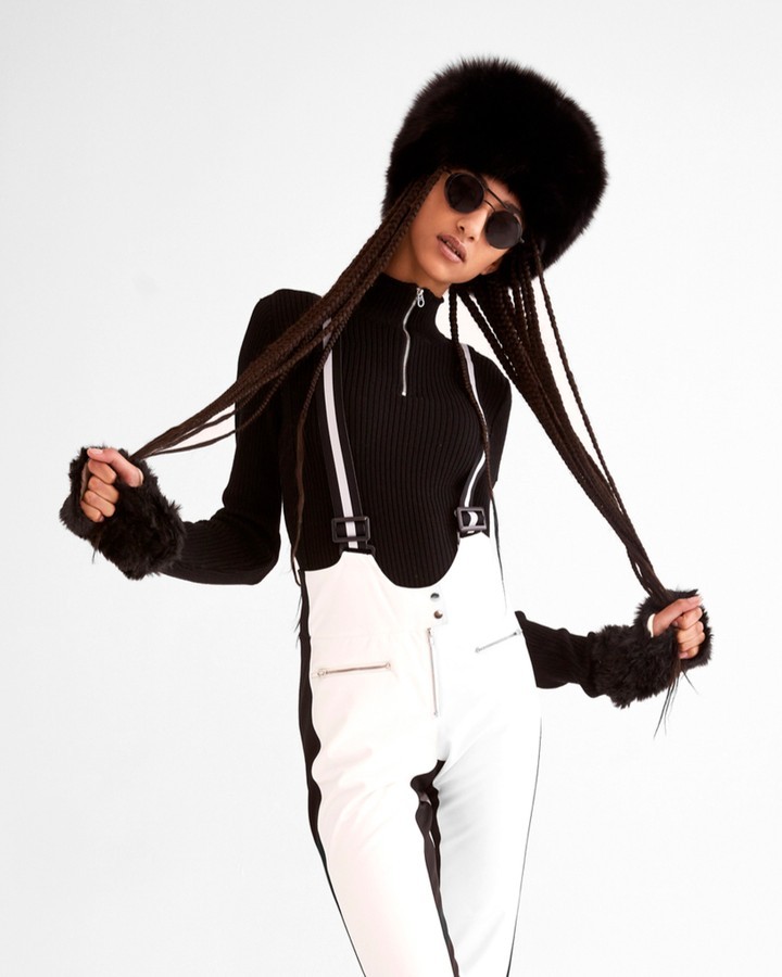 Woman in black turtleneck and white overalls wearing a furry hat and sunglasses, posing against a white backdrop in a studio setting.