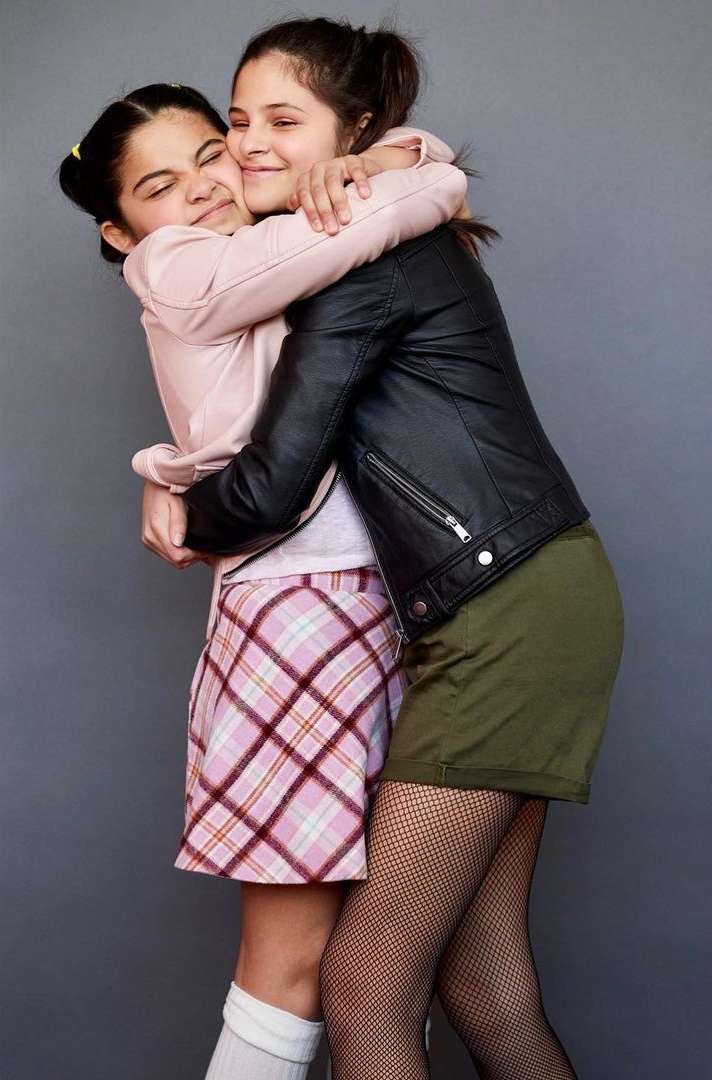 Two girls embracing tightly indoors, wearing contrasting outfits - one in pinks and the other in darker greens and black, smiling.