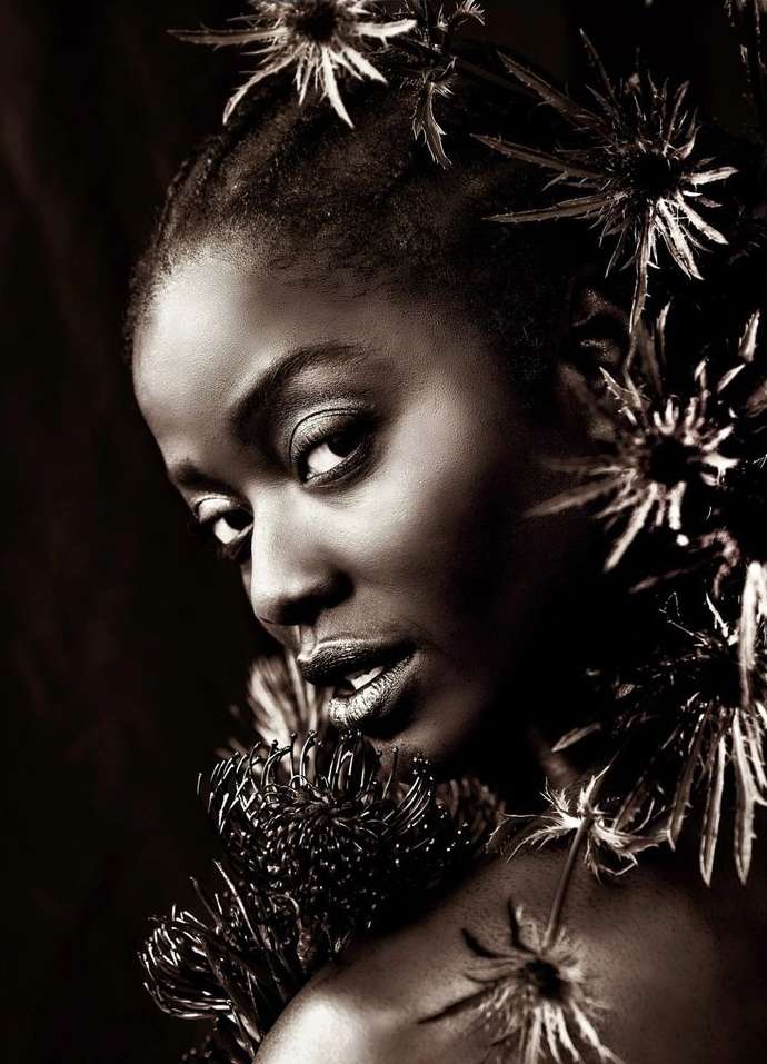Portrait of a Black woman with dark skin, wearing dark lipstick and surrounded by spiky dark-colored flowers on a dark background.