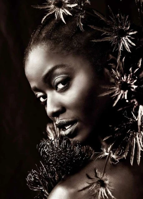 Portrait of a Black woman with dark skin, wearing dark lipstick and surrounded by spiky dark-colored flowers on a dark background.