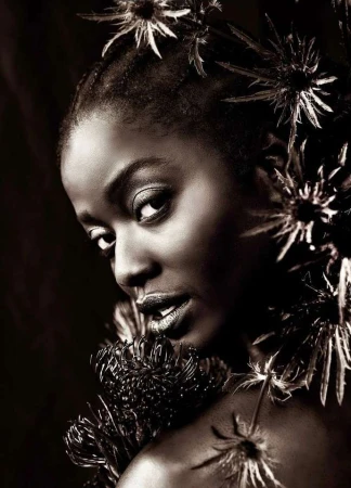 Portrait of a Black woman with dark skin, wearing dark lipstick and surrounded by spiky dark-colored flowers on a dark background.