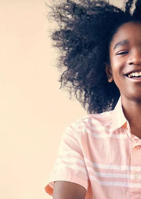Close-up portrait of a smiling, dark-skinned child with natural afro hair, wearing a peach-colored striped shirt against a neutral backdrop.