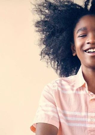 Close-up portrait of a smiling, dark-skinned child with natural afro hair, wearing a peach-colored striped shirt against a neutral backdrop.