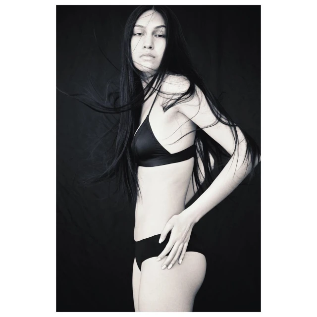 Monochrome portrait of a woman with long black hair wearing a bra and panties, striking a pose against a solid black background.