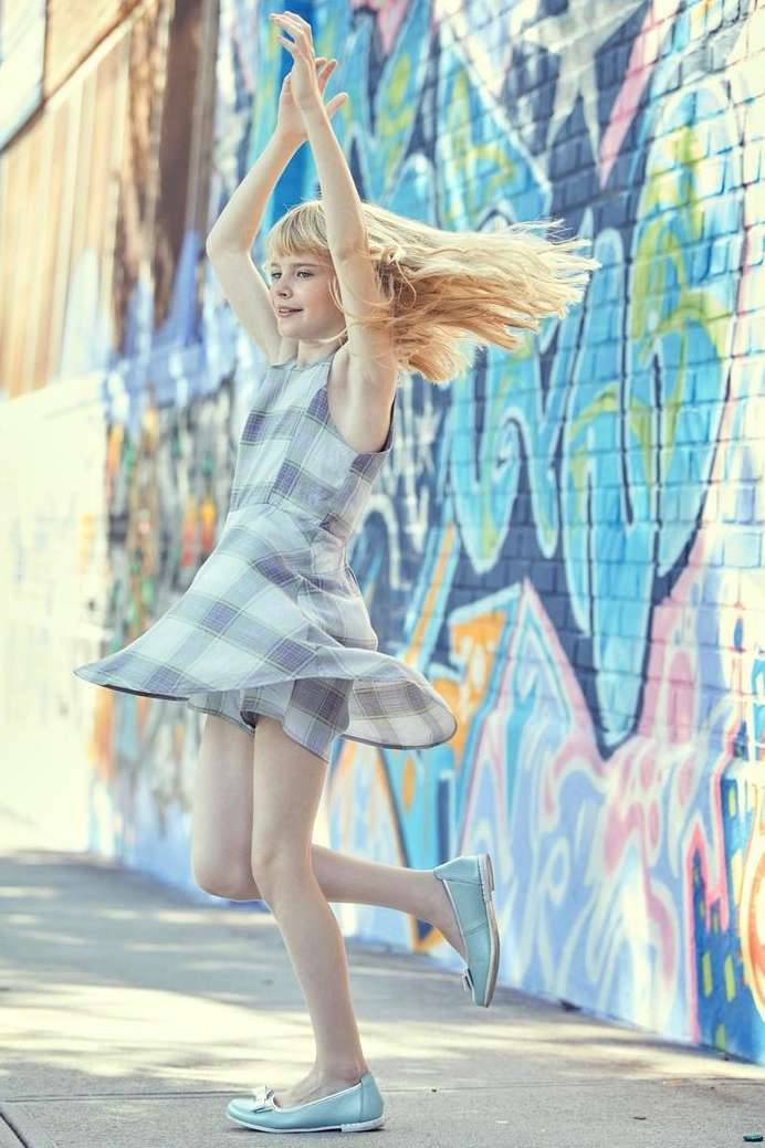 Young girl twirling in a plaid dress, arms raised in front of a graffiti wall. Blonde hair, light skin, ballet flats, outdoor setting.