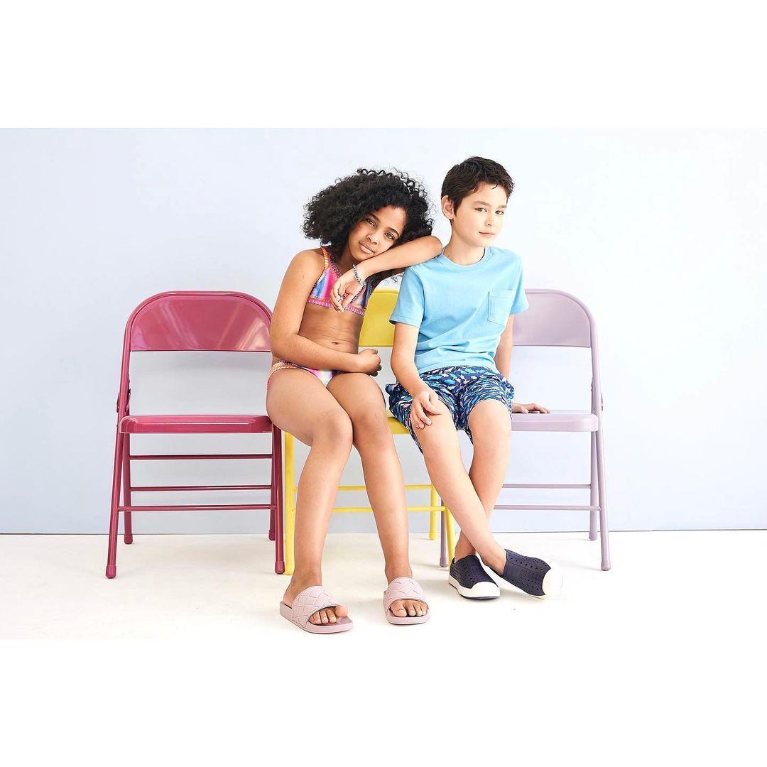 Two kids, a girl and a boy, sit together on metal folding chairs in a studio setting, wearing swimwear and casual clothing.