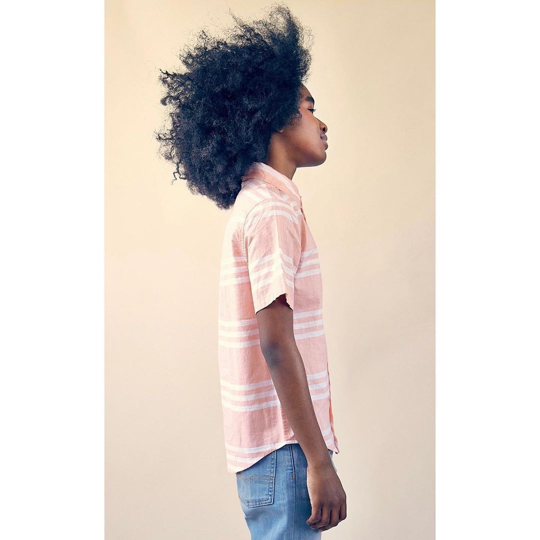 Profile portrait of African-American male in pink striped shirt and blue jeans standing against a beige background in an indoor setting.