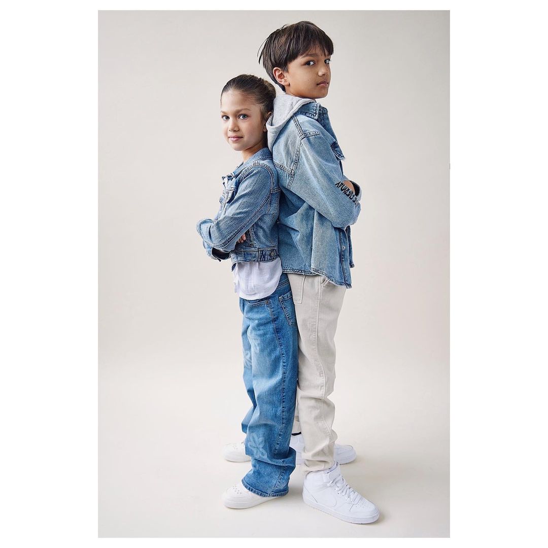 Portrait of a boy and a girl standing back-to-back in denim jackets and jeans against a neutral background in a studio setting.
