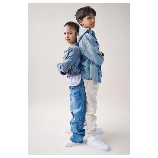 Portrait of a boy and a girl standing back-to-back in denim jackets and jeans against a neutral background in a studio setting.