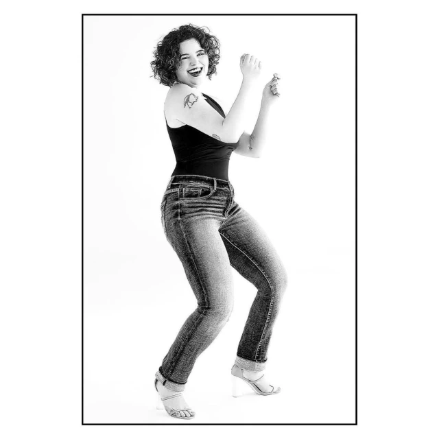 Black and white full shot of an adult female with curly hair, jeans, tank top, and high heels dancing in a white photo studio.