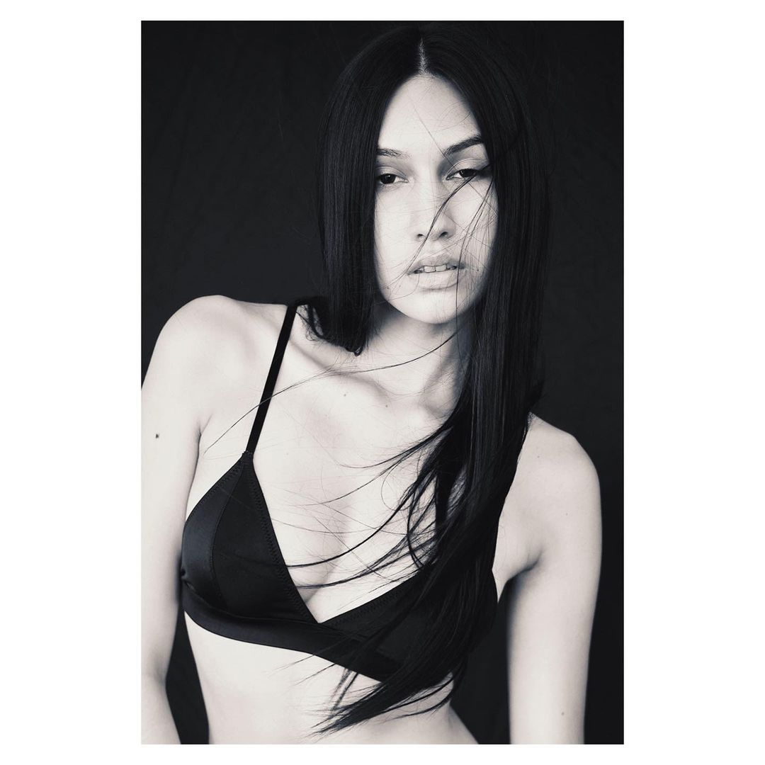 Black and white portrait of a woman with long dark hair wearing a dark bra top against a dark background