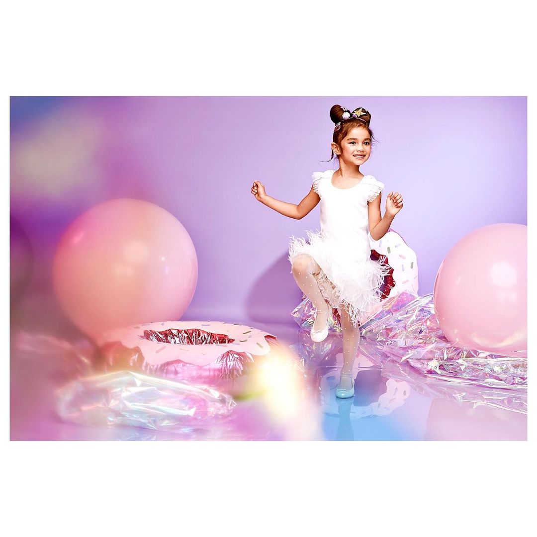 Adorable young girl in a white tutu dress poses playfully among giant pink balloons and shiny donut prop against a lavender background.
