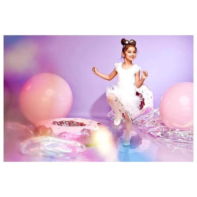 Adorable young girl in a white tutu dress poses playfully among giant pink balloons and shiny donut prop against a lavender background.