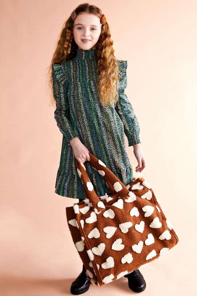 Young girl with long red hair poses in a green patterned dress with a heart-patterned tote bag, creating a charming fashion portrait.