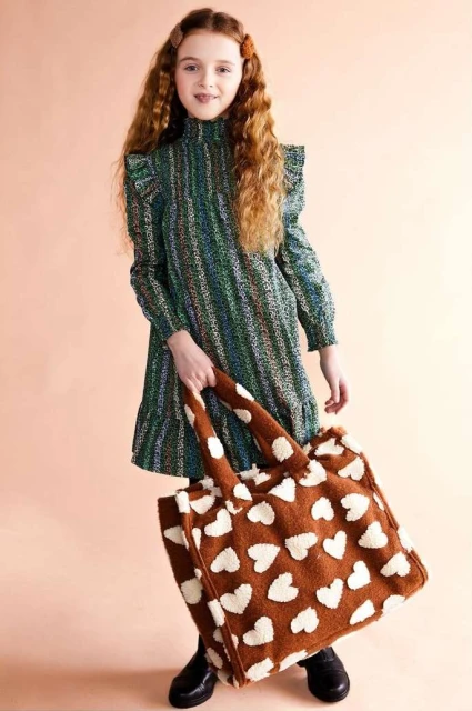Young girl with long red hair poses in a green patterned dress with a heart-patterned tote bag, creating a charming fashion portrait.