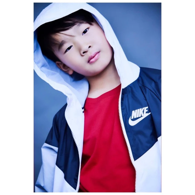 Portrait of an Asian child with fair skin wearing a red t-shirt and blue and white hooded windbreaker jacket in a studio setting.