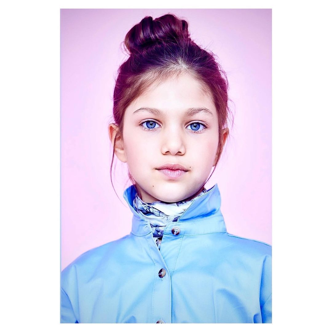 Portrait of a young girl with blue eyes and auburn hair in a high bun, wearing a blue jacket against a light pink backdrop.