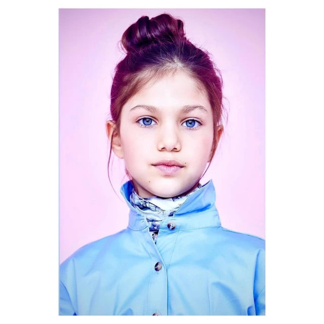 Portrait of a young girl with blue eyes and auburn hair in a high bun, wearing a blue jacket against a light pink backdrop.