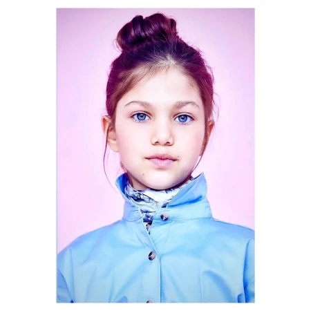 Portrait of a young girl with blue eyes and auburn hair in a high bun, wearing a blue jacket against a light pink backdrop.