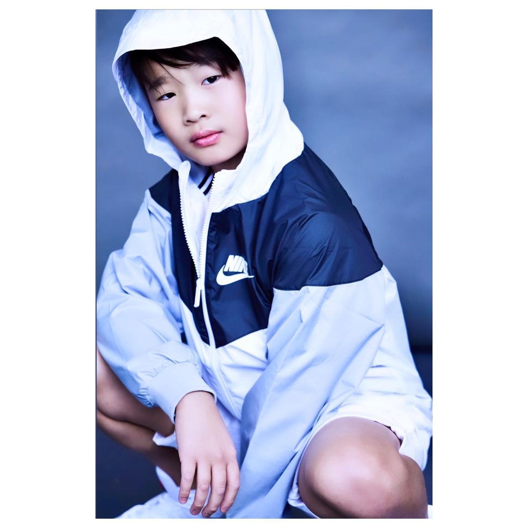 A young child wearing a Nike windbreaker jacket with a hood poses crouching indoors against a grey background.