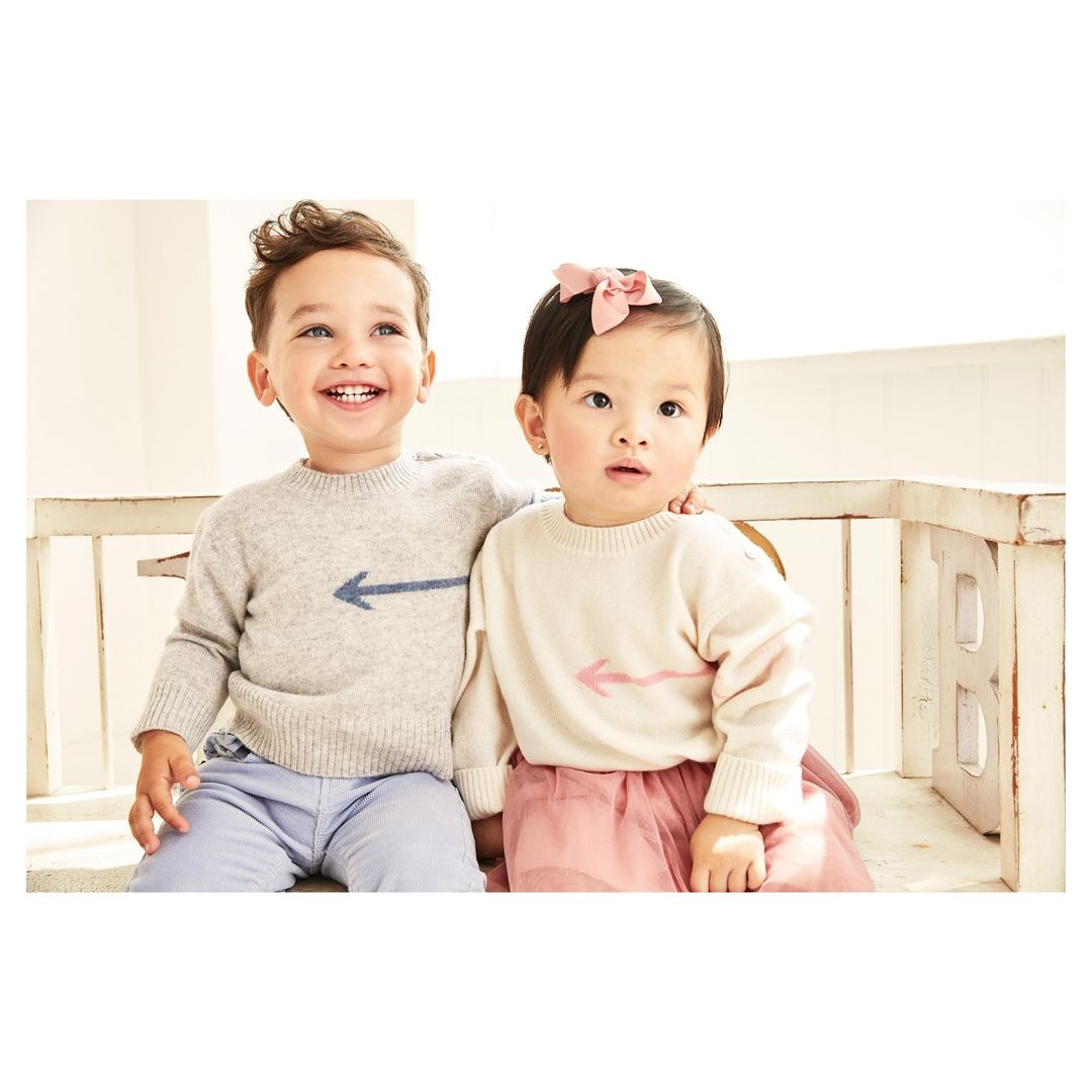 Two toddlers, a boy and a girl, wear matching sweaters with arrow graphics, posing together indoors against a light backdrop and white bench.