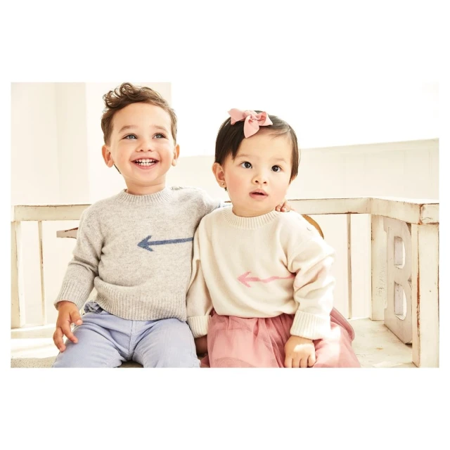 Two toddlers, a boy and a girl, wear matching sweaters with arrow graphics, posing together indoors against a light backdrop and white bench.