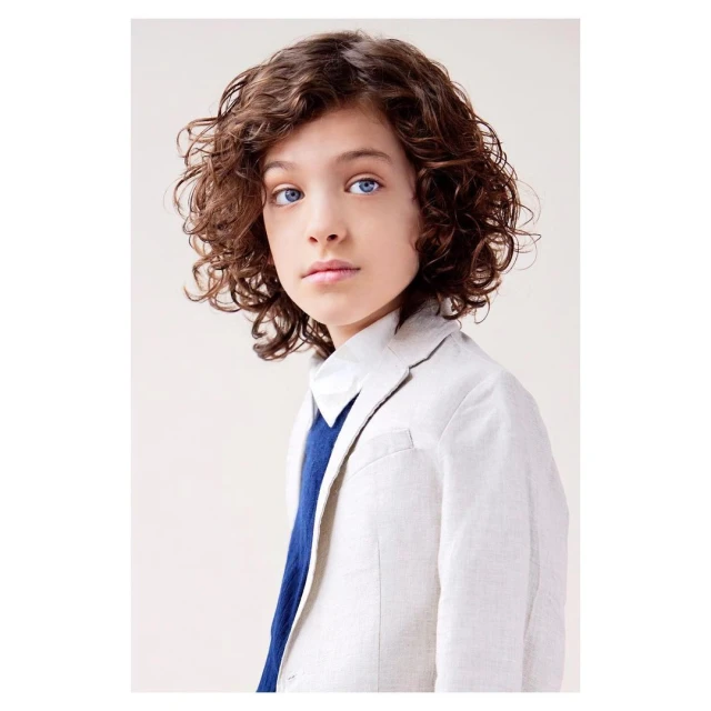 Portrait of a boy with curly brown hair wearing a linen blazer, set against a neutral backdrop in a studio setting.