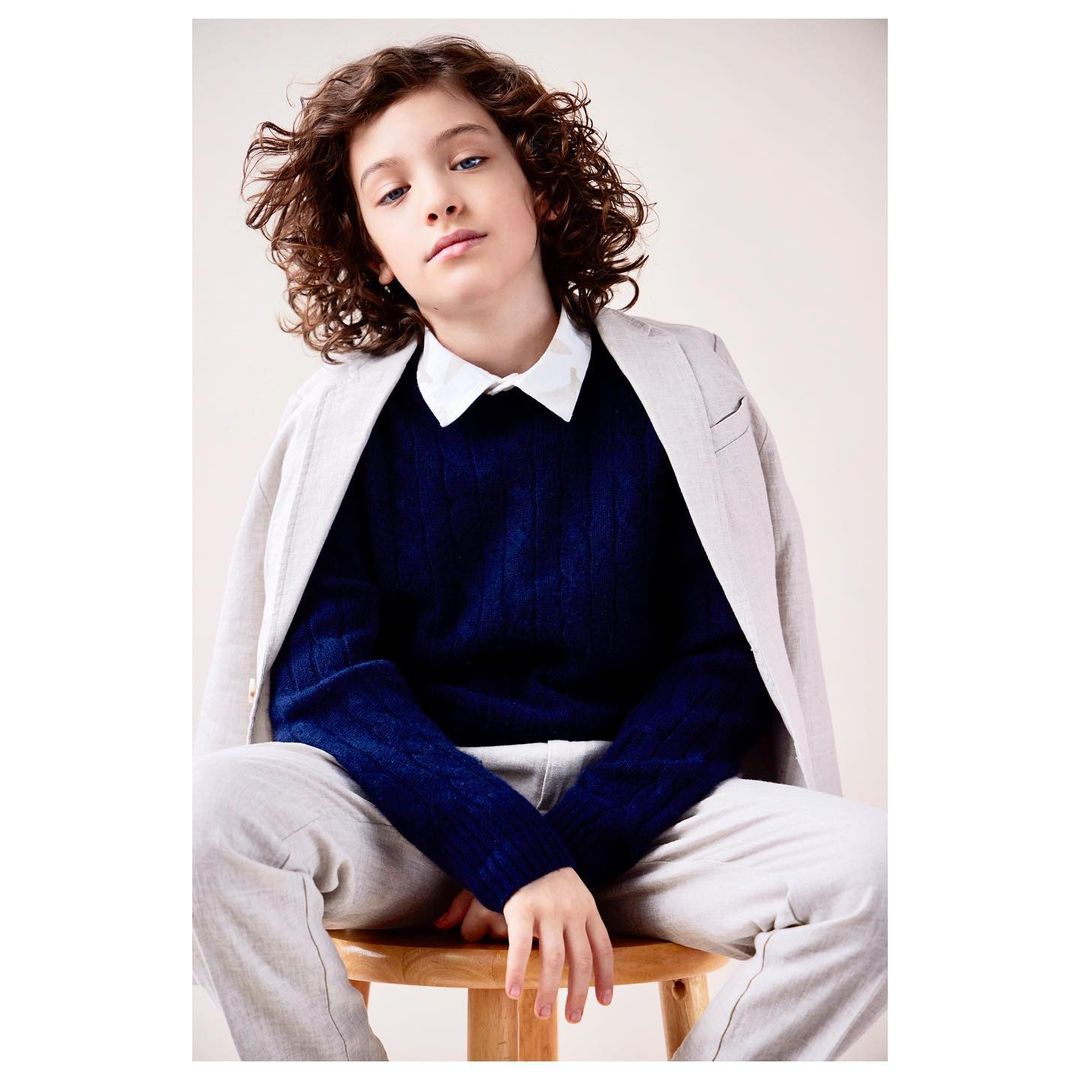 Portrait of a stylish young boy with curly hair wearing a blazer and blue sweater, seated on a wooden stool against a neutral backdrop.