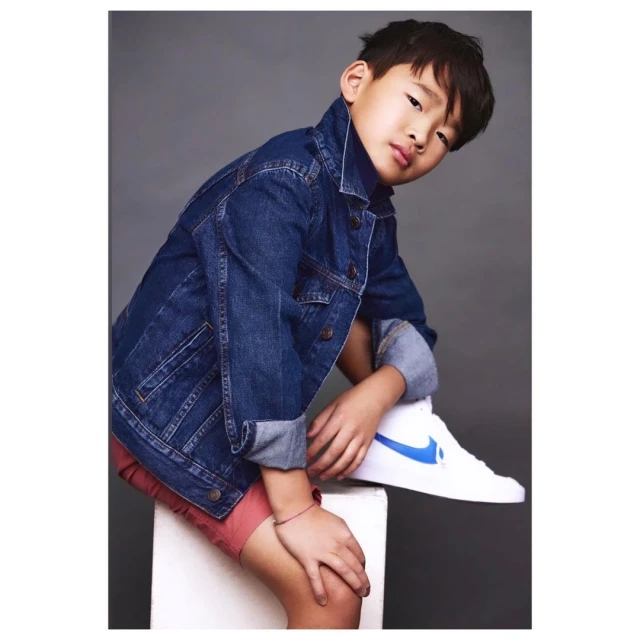 Young boy in a denim jacket and red shorts sitting on a white cube with a white sneaker and blue Nike swoosh, posing in a photo studio.